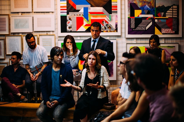 A group of people are gathered in a room with colorful abstract art on the walls. The central figure appears to be speaking or expressing a point, while others listen attentively. The setting suggests a casual yet focused discussion or meeting.