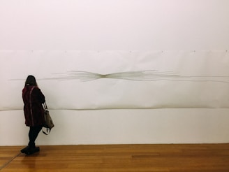 A person stands in front of a minimalist art piece on a gallery wall. The artwork features linear, abstract designs that converge at the center, resembling a vortex or knot. The individual, wearing a dark coat and carrying a shoulder bag, seems to be observing the piece closely. The gallery floor is wooden, adding warmth to the neutral tones of the wall and art.