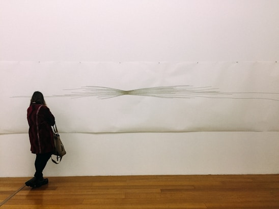 A person stands in front of a minimalist art piece on a gallery wall. The artwork features linear, abstract designs that converge at the center, resembling a vortex or knot. The individual, wearing a dark coat and carrying a shoulder bag, seems to be observing the piece closely. The gallery floor is wooden, adding warmth to the neutral tones of the wall and art.