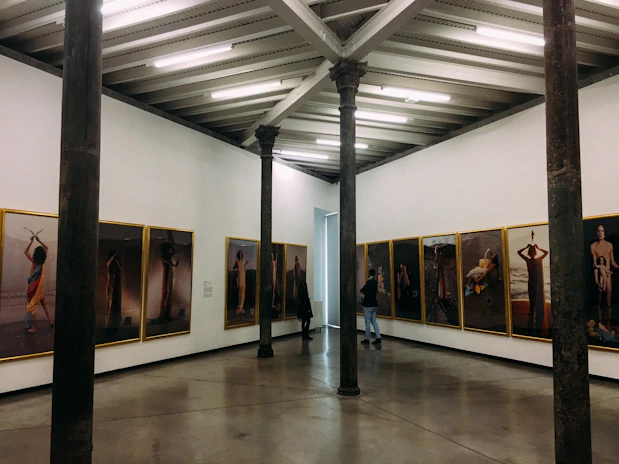 An art gallery with framed photographs hung on the walls in a bright room. The pictures display various human figures and scenes. The gallery features exposed beams on the ceiling and tall, dark columns within the space. Two people are present, observing the art.