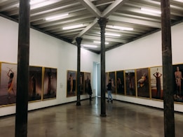An art gallery with framed photographs hung on the walls in a bright room. The pictures display various human figures and scenes. The gallery features exposed beams on the ceiling and tall, dark columns within the space. Two people are present, observing the art.