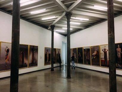 An art gallery with framed photographs hung on the walls in a bright room. The pictures display various human figures and scenes. The gallery features exposed beams on the ceiling and tall, dark columns within the space. Two people are present, observing the art.
