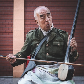 An elderly man is playing a stringed musical instrument, resembling an erhu, against a brick wall background. He is wearing a green military-style jacket and has a white apron on his lap. His expression appears focused and contemplative.