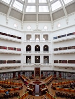 The image depicts a grand library with a large, open reading area at the center. The architecture features a stunning dome ceiling with glass panels allowing natural light to flood the interior. Surrounding the reading area are several tiers of book-lined balconies, accessed by intricate staircases and railings. The space is elegantly decorated in white with ornate detailing, and several people are seated at wooden desks, reading under classic green lamps.