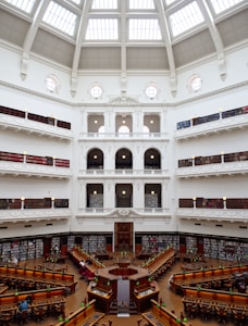 The image depicts a grand library with a large, open reading area at the center. The architecture features a stunning dome ceiling with glass panels allowing natural light to flood the interior. Surrounding the reading area are several tiers of book-lined balconies, accessed by intricate staircases and railings. The space is elegantly decorated in white with ornate detailing, and several people are seated at wooden desks, reading under classic green lamps.