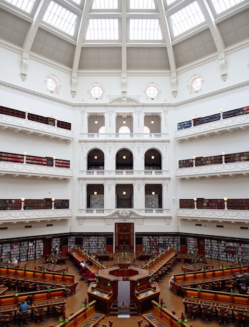 The image depicts a grand library with a large, open reading area at the center. The architecture features a stunning dome ceiling with glass panels allowing natural light to flood the interior. Surrounding the reading area are several tiers of book-lined balconies, accessed by intricate staircases and railings. The space is elegantly decorated in white with ornate detailing, and several people are seated at wooden desks, reading under classic green lamps.