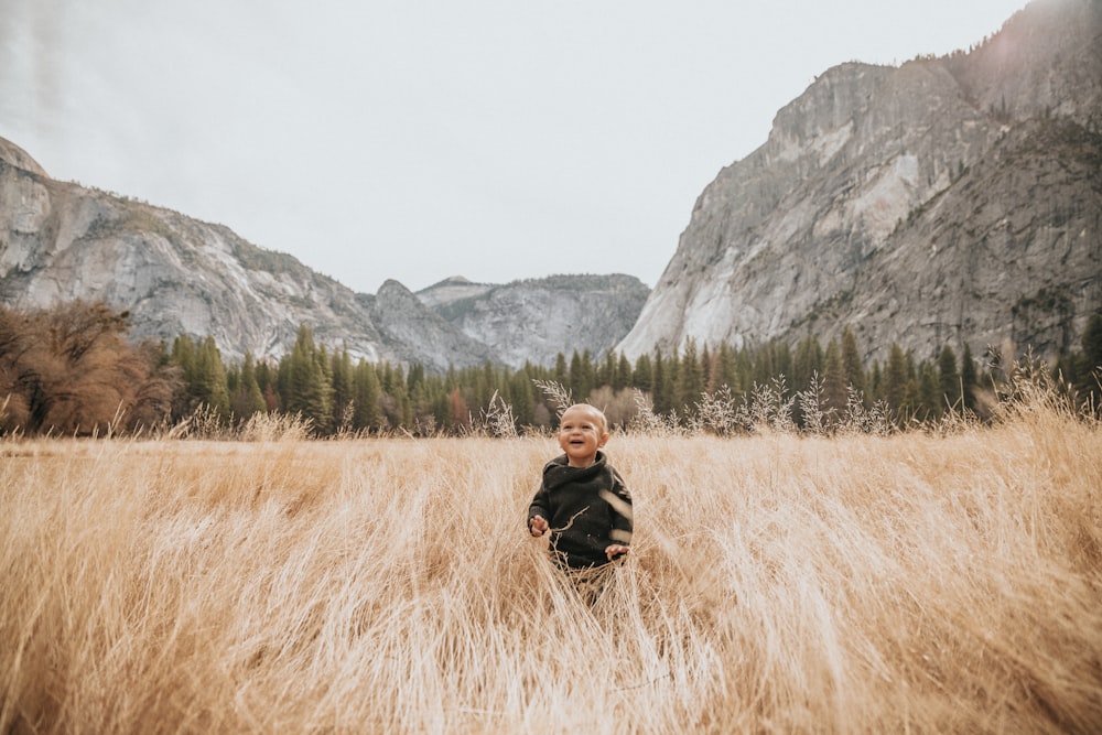 boy in black jacket stand on brown grass near mountain
