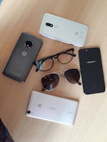 The image displays several smartphones, glasses, and sunglasses arranged on a wooden surface. The phones include brands like Motorola, Oppo, and Vivo.