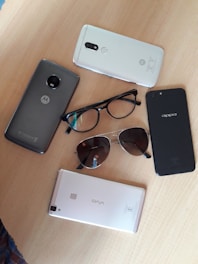 The image displays several smartphones, glasses, and sunglasses arranged on a wooden surface. The phones include brands like Motorola, Oppo, and Vivo.