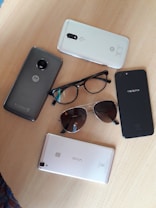 The image displays several smartphones, glasses, and sunglasses arranged on a wooden surface. The phones include brands like Motorola, Oppo, and Vivo.