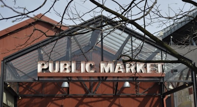 A large sign reading 'Public Market' is affixed to the facade of a building with a sloped glass and metal roof. Leafless branches of a tree are in the foreground, slightly obscuring the view of the sign. The architecture suggests an industrial or warehouse style.