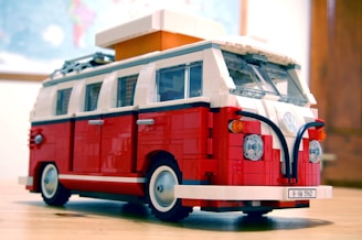 A detailed LEGO model of a classic Volkswagen camper van in red and white, featuring detailed exterior elements like windows, headlights, and a rooftop box. It is placed on a wooden surface with a blurred background, suggesting an indoor environment.