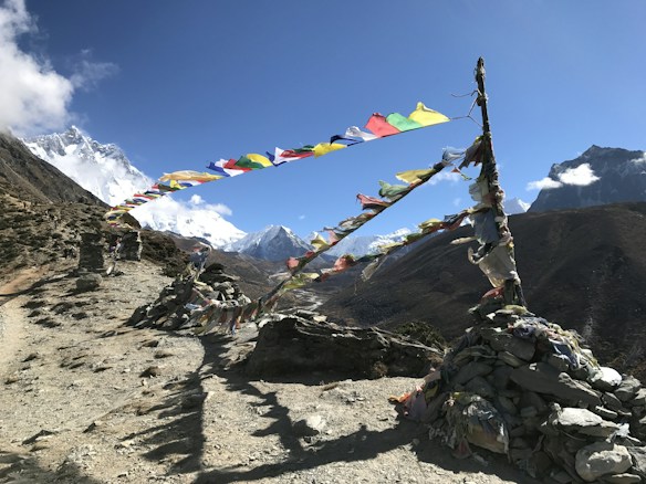 A mountainous landscape featuring colorful prayer flags strung across stone cairns. Snow-capped peaks rise in the background, while a clear blue sky dominates the scene. The path runs alongside the prayer flags, offering a breathtaking view of the rugged terrain.