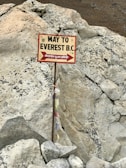 A signpost is directing the way to Everest Base Camp (B.C.), with text in English and Nepali. The sign is mounted on a metal pole with some colorful prayer flags attached to it. The background consists of large, rough, rocky terrain.