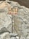 A signpost is directing the way to Everest Base Camp (B.C.), with text in English and Nepali. The sign is mounted on a metal pole with some colorful prayer flags attached to it. The background consists of large, rough, rocky terrain.