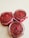 Three glass jars filled with a textured red substance, possibly a homemade jam or spread, are arranged on a light surface. Each jar is topped with a single red berry and is decorated with a red ribbon around the rim.