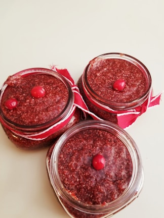 Three glass jars filled with a textured red substance, possibly a homemade jam or spread, are arranged on a light surface. Each jar is topped with a single red berry and is decorated with a red ribbon around the rim.