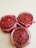 Three glass jars filled with a textured red substance, possibly a homemade jam or spread, are arranged on a light surface. Each jar is topped with a single red berry and is decorated with a red ribbon around the rim.