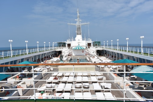 An open deck of a cruise ship features numerous sun loungers under white towels, a pool in the center, and striped umbrellas providing shade. The ship's railings and the vast ocean are visible in the background.