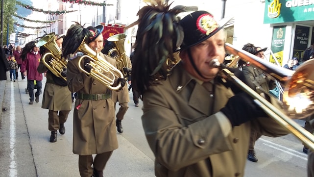 A brass band is marching down a street, with several musicians playing instruments like trumpets and trombones. They are dressed in uniform coats and feathered hats. Behind them, a crowd of people is watching the parade, and there are shops and decorations along the street.