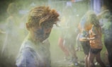 A group of people engaging in a colorful powder activity, with vibrant hues covering their clothing and bodies. The atmosphere is filled with dust and laughter, suggesting a festive and playful setting.