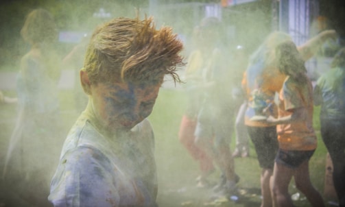 A group of people engaging in a colorful powder activity, with vibrant hues covering their clothing and bodies. The atmosphere is filled with dust and laughter, suggesting a festive and playful setting.
