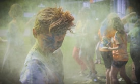 A group of people engaging in a colorful powder activity, with vibrant hues covering their clothing and bodies. The atmosphere is filled with dust and laughter, suggesting a festive and playful setting.