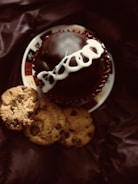 A chocolate-covered cupcake with white icing on top, placed on a decorative plate, accompanied by three chocolate chip cookies on a dark-colored fabric background.