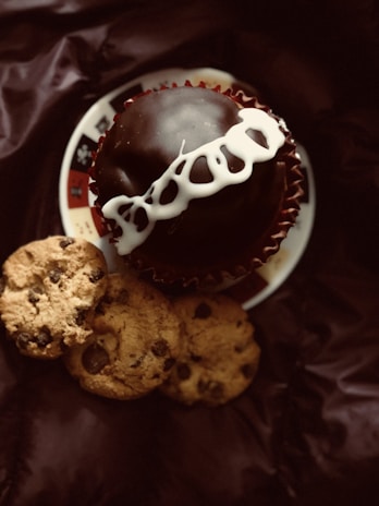 A chocolate-covered cupcake with white icing on top, placed on a decorative plate, accompanied by three chocolate chip cookies on a dark-colored fabric background.