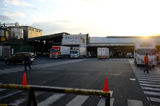 A busy industrial area features several large trucks parked in front of a warehouse. Two people are walking across the street in different directions. The sun sets in the background, casting a warm glow over the scene. Traffic cones and barriers are situated in the foreground, adding elements of safety and organization.