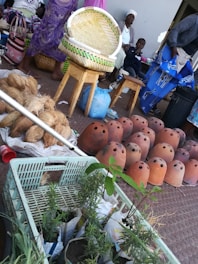 A bustling market scene with people and various items. There are several clay pots with holes, a basket placed on a wooden stool, coconuts, and small potted plants in the foreground. People are wearing colorful attire and sitting or standing around, indicating a lively environment.