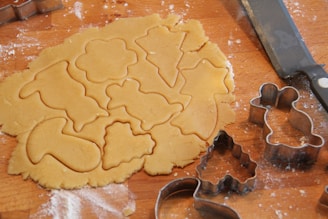 A rolled-out piece of dough with various shapes cut out using cookie cutters, including stars, trees, and animal figures. The background shows a wooden surface with scattered flour and a knife.