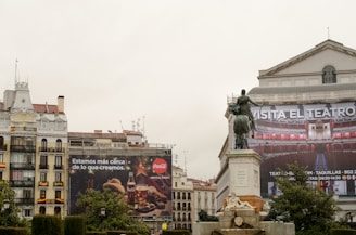 A cityscape featuring a large advertisement on an old building with a Coca-Cola logo and a Spanish phrase. There's also a large banner encouraging people to visit the theater, with an image of an ornate theater interior. A statue of a person on horseback is prominently in front of the building, with a fountain at the base.