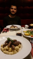 A person sitting at a dining table with two plates of food in the foreground. One dish contains gnocchi topped with grilled slices of meat and garnished with herbs. A glass of red wine is next to it. In the background, there is a second plate with a salad featuring sliced yellow peppers and tomatoes.