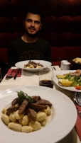 A person sitting at a dining table with two plates of food in the foreground. One dish contains gnocchi topped with grilled slices of meat and garnished with herbs. A glass of red wine is next to it. In the background, there is a second plate with a salad featuring sliced yellow peppers and tomatoes.