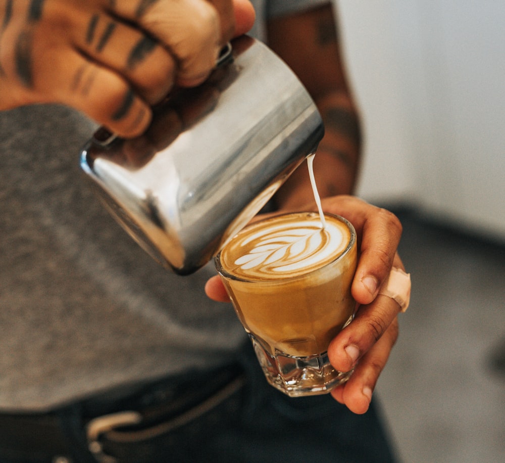 man holding clear drinking glass with coffee and pouring with milk