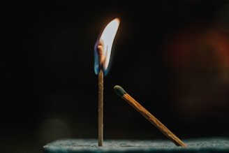 A matchstick is upright with a flame burning at its tip, casting a warm glow. Another unlit matchstick leans diagonally against a surface, positioned towards the flame. The background is dark, emphasizing the brightness and colors of the flame.