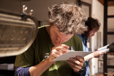 A person with disheveled hair is closely examining a piece of paper using a small magnifying lens. Wearing a green shirt, they appear focused in a well-lit room with another person in the background also looking at something. The scene suggests an environment of detailed inspection or analysis.