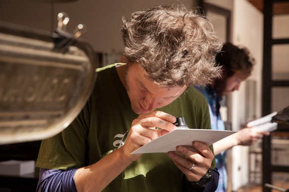 A person with disheveled hair is closely examining a piece of paper using a small magnifying lens. Wearing a green shirt, they appear focused in a well-lit room with another person in the background also looking at something. The scene suggests an environment of detailed inspection or analysis.