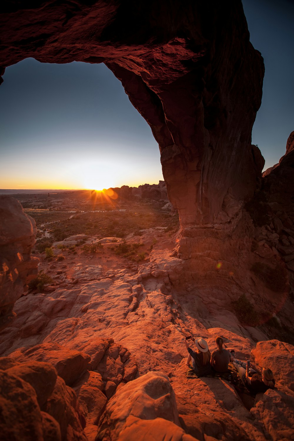 two people sitting on rock below arch facing sun