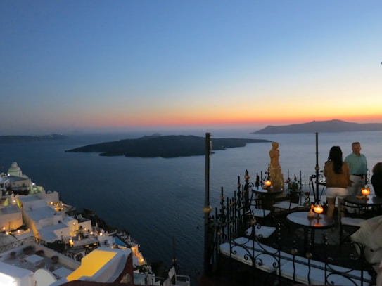 A stunning panoramic view of the ocean at sunset with a vibrant gradient of warm hues in the sky. White buildings perched on a cliffside, characteristic of Santorini, provide a picturesque backdrop. Several tables with lit candles are arranged on an open terrace, with a couple of individuals enjoying the view.