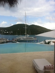 A serene coastal scene featuring a luxurious infinity pool overlooking a calm body of water. A sailboat is moored nearby, with a backdrop of lush, green hills under a partly cloudy sky. There is a sun lounger beside the pool, suggesting relaxation.