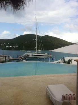 A serene coastal scene featuring a luxurious infinity pool overlooking a calm body of water. A sailboat is moored nearby, with a backdrop of lush, green hills under a partly cloudy sky. There is a sun lounger beside the pool, suggesting relaxation.