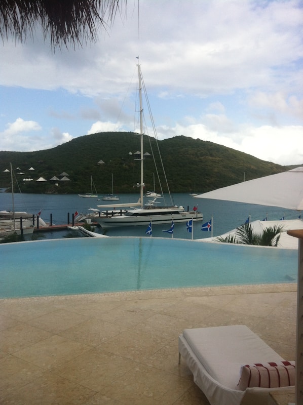 A serene coastal scene featuring a luxurious infinity pool overlooking a calm body of water. A sailboat is moored nearby, with a backdrop of lush, green hills under a partly cloudy sky. There is a sun lounger beside the pool, suggesting relaxation.