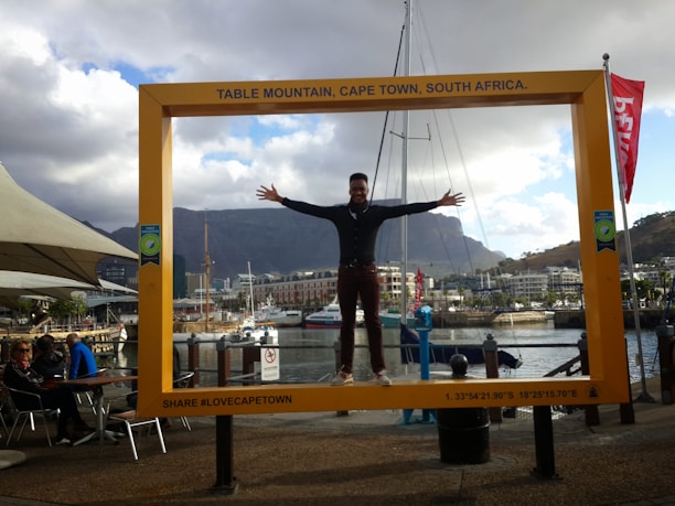 A person is standing with arms outstretched inside a large, yellow picture frame that has 'Table Mountain, Cape Town, South Africa' written on top. In the background, there are people seated at a cafe by the waterfront, with boats docked on the water and a mountain range visible. The sky is partly cloudy.