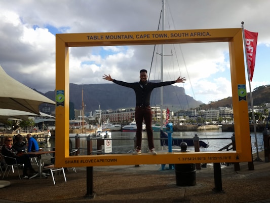 A person is standing with arms outstretched inside a large, yellow picture frame that has 'Table Mountain, Cape Town, South Africa' written on top. In the background, there are people seated at a cafe by the waterfront, with boats docked on the water and a mountain range visible. The sky is partly cloudy.