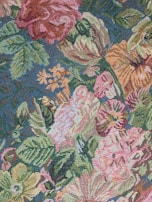 An intricately woven tapestry features a vibrant floral pattern with large blooms including roses and other various flowers. The design includes lush green leaves intertwined with colorful blossoms on a textured, fabric-like surface.