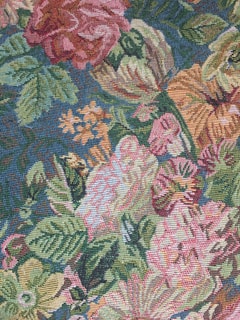 An intricately woven tapestry features a vibrant floral pattern with large blooms including roses and other various flowers. The design includes lush green leaves intertwined with colorful blossoms on a textured, fabric-like surface.