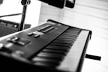 A black and white photograph of an electronic keyboard, showcasing the keys and controls in a close-up perspective. The keyboard is situated indoors, and a blurred amplifier or speaker is visible in the background.