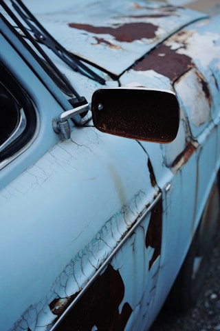A close-up view of an old car with significant rust damage and peeling paint. The surface appears weathered, with various shades of blue and patches of reddish-brown rust. The side mirror is visible, along with part of the windshield and wipers.